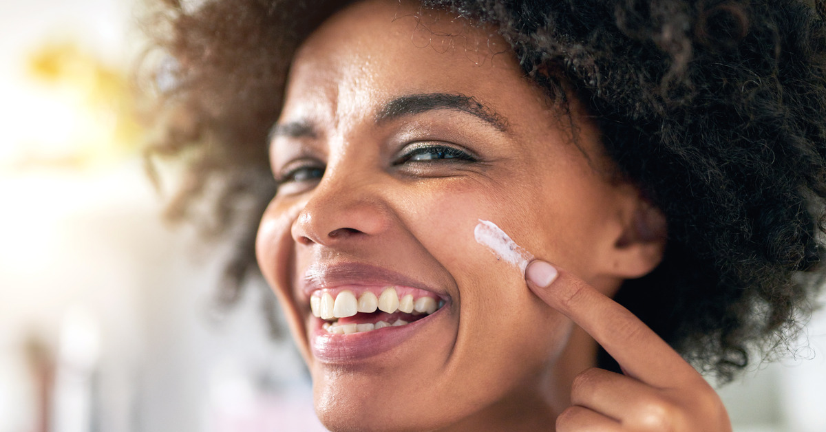 Portrait shot of a Black woman smiling as she smears a line of sunscreen across her cheek with her finger.